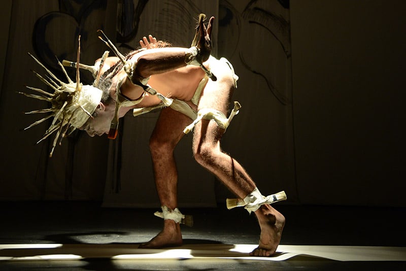 Photo of a dancer in a black empty stage space, he is strongly brightly lit, almost unclothed with an abstract costume and headgear in white, like spikes, he has his upper body bent forward. White masking tape is wrapped around his joints and wide brushes are attached to it.