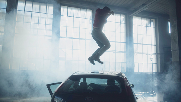  A dark blue car in an industrial hall. The car is surrounded by white smoke and is backlit by a window front. A person on the roof of the car jumps into the air. The face is unrecognisable. The person is wearing jeans, a red bomber jacket and boots. The arms are raised in the air.