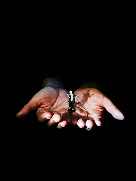 Two hands emerge from the darkness. In the middle of the two palms stands a cardboard figure, screaming with raised hands.