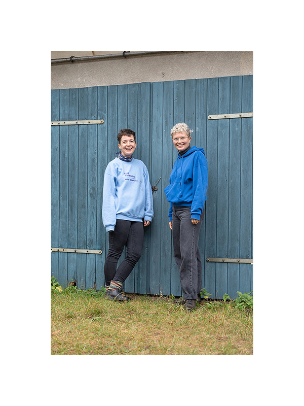 Zwei Menschen stehen nebeneinander vor einer blauen Holztür auf einer Wiese. Beide tragen blaue Pullover, kurze Haare und sind weiß. Sie lächeln in die Kamera.