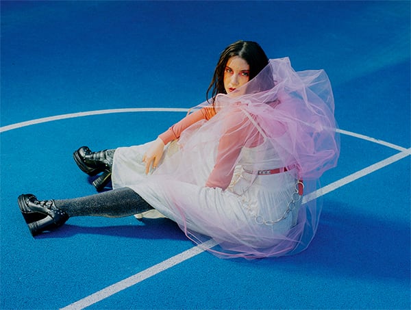  A woman sits on a bright blue basketball court, leaning slightly. She wears an eclectic outfit: a flowy white dress with pink tulle, black platform shoes, sparkly grey tights, and bold makeup. The scene feels whimsical and avant-garde, mixing soft fabrics with edgy footwear.