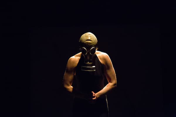 Foto eines Menschen mit Gasmaske und schwarzer Lederschürze, mit freien Armen auf der Bühne von oben beleuchtet