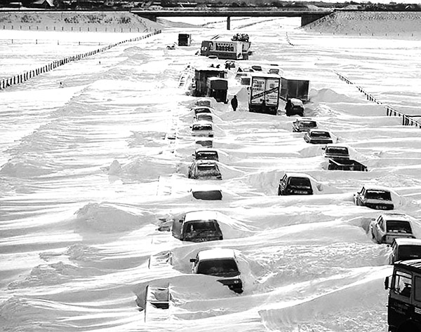 Black and white photograph of a snowed up highway with trucks and cars buried in snow in wide landscape