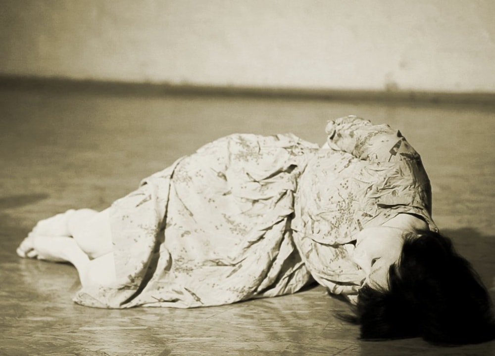 Black and white photograph of a young woman in a dress lying on her side on the stage floor.