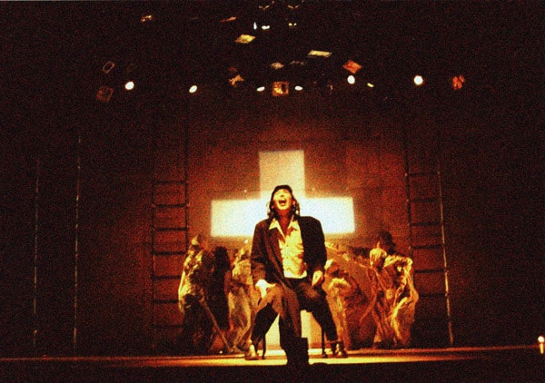 Foto des Bühnengeschehens ein Tänzerim Vordergrund, ein leuchtendes Kreuz im Hintergrund