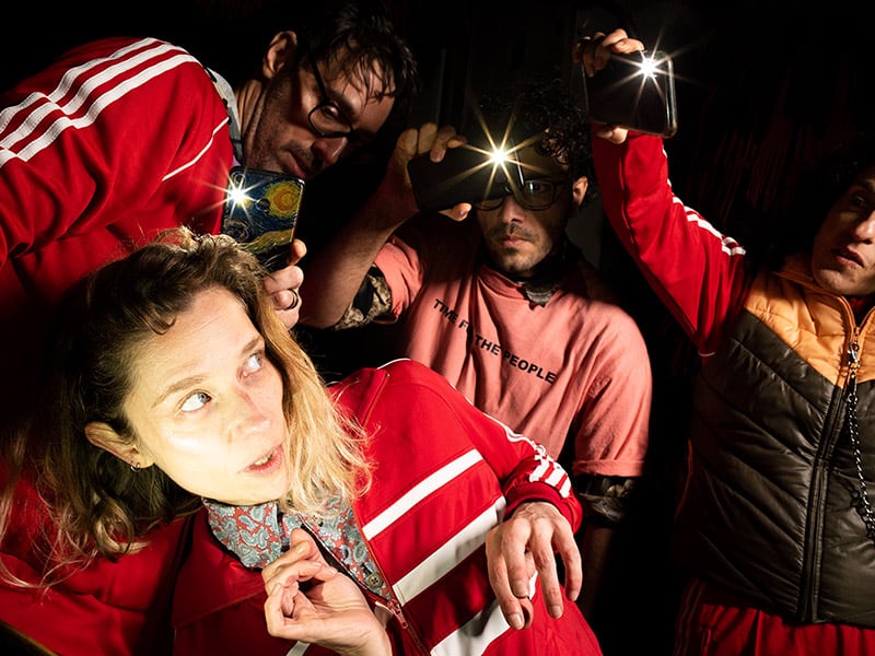 A dynamic shot in which three people are photographing a middle-aged woman with cell phones. She is in the foreground and in the center of the picture and is strongly lit. All of them are wearing red track jackets. The flash of the cell phones can be seen.