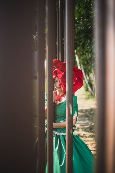 Foto einer Frau mit roter Kopfbedeckung unt grünem Kleid draußen hinter Gitterstäben