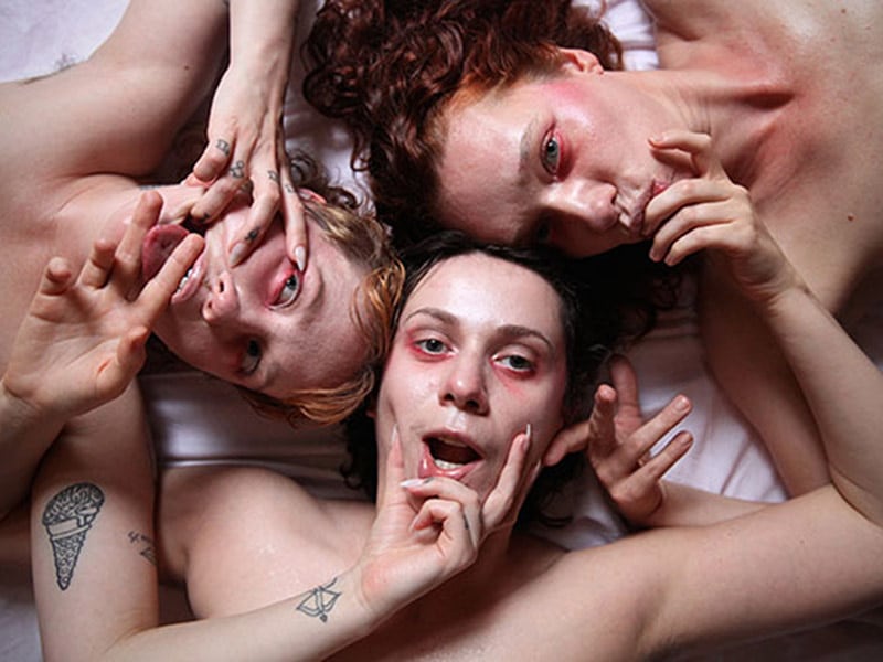  Close up of three young people with naked torsos and tattoos, and red eyeliner. They touch each other's mouths with their hands and look directly into the camera.