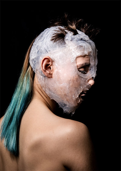 Close-up of a woman from behind. Her face looks back, she wears a kind of mask made of transparent white plastic, on her shoulder falls a ponytail of blue-green hair.