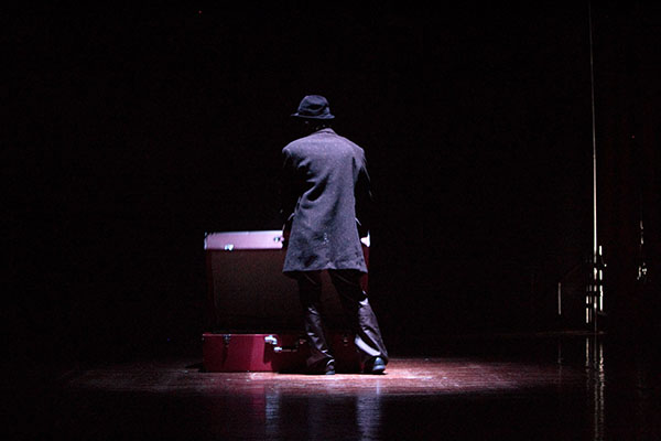 Fotografie einer Person von hinten im Bühnenraum in Mantel mit Hut und mit Koffern