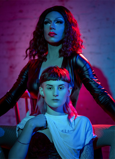 A photo of two female read nonbinary people in blue, red spotlight. 