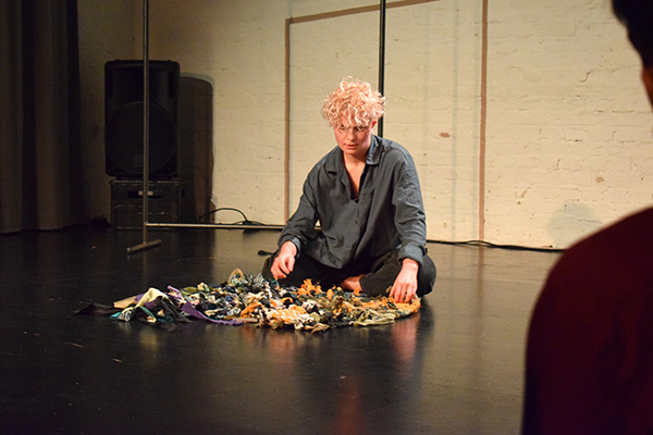 Eine Person mit kurzen, lockigen Haaren sitzt auf dem Boden - vor sich ein Textil, das aus verschiedenfarbigen Stoffen gewebt ist. Die Person hat den Blick auf das Textil gesenkt und berührt es sanft mit den Händen. 