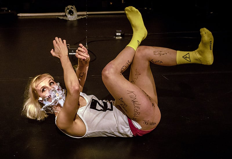 A woman lies on her back with her legs and arms raised. Her mouth is covered by a plastic mask with letters cut out of newspapers.
