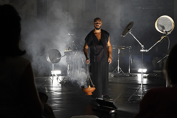 A man in a sleeveless black outfit stands on a dark, smoky stage, holding a clay incense burner that releases smoke. Around him are percussion instruments and microphones under soft white lighting, while audience members watch from the shadows.