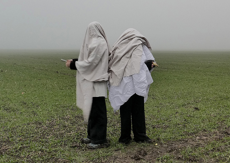 Two people are standing with their backs to each other in a misty, wide field. They have thrown over sheets, only their legs and hands are visible. They are each peeling an unrecognisable fruit. Their silhouettes are reminiscent of those of ghosts.