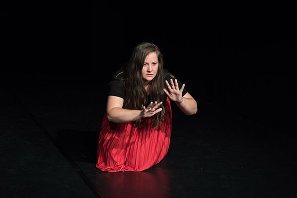 Im Zentrum des Fotos ist eine junge Schauspielerin in einem roten glänzenden Rock im schwarzen Bühnenraum kniend, gestikulierend, sprechend Richtung Zuschauer*innen gerichtet.