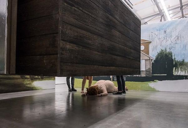 Photography of an exhibition space with room installation, a painting in the background and a woman in a fatsuit lying on her stomach, her face to the floor under a floating wooden object.