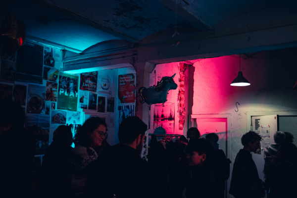 Ein belebter Innenraum mit mehreren Menschen, die im Halbdunkel zusammenstehen und sich unterhalten. An den Wänden hängen viele Poster, beleuchtet von blauem und pinkem Neonlicht, daneben eine Hängelampe und ein Einhorn-Ballon.