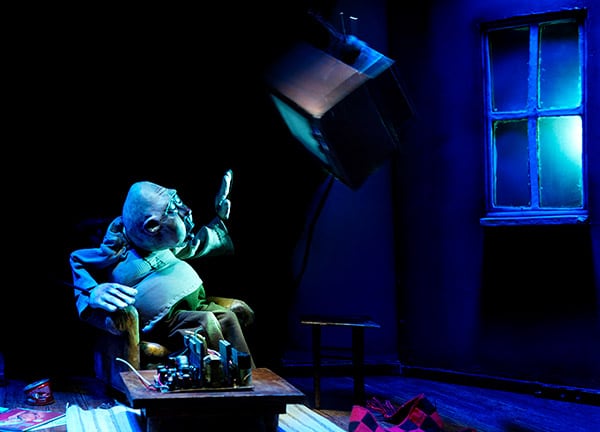 Photography of a mannequin in a small living room setting depicting an elderly balding pot-bellied man sitting on an armchair, he has his hand raised as the light falling through a small window dazzles him. On the table in front of him lies an object with cables on which he seemed to be building.
