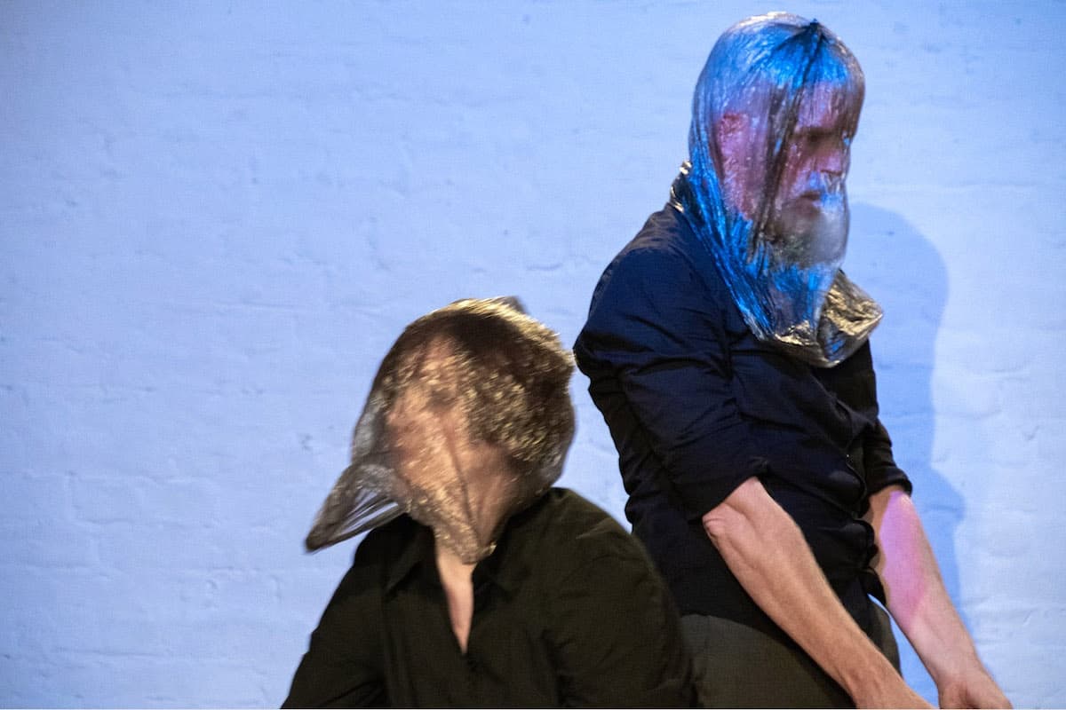 Photography of two people with gray transparent plastic bags over their heads in black clothes in motion in front of a white wall. Blue spotlight reflects on the bags.