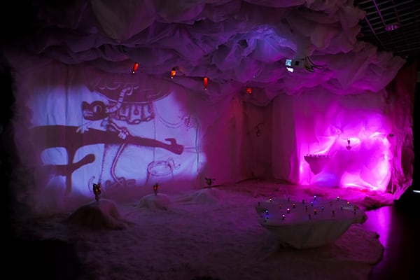 An immersive art installation featuring soft white fabric covering the walls, ceiling, and floor, illuminated by purple and pink lights. A cartoon-style image is projected on one wall, while small colorful lights glow on the ground.